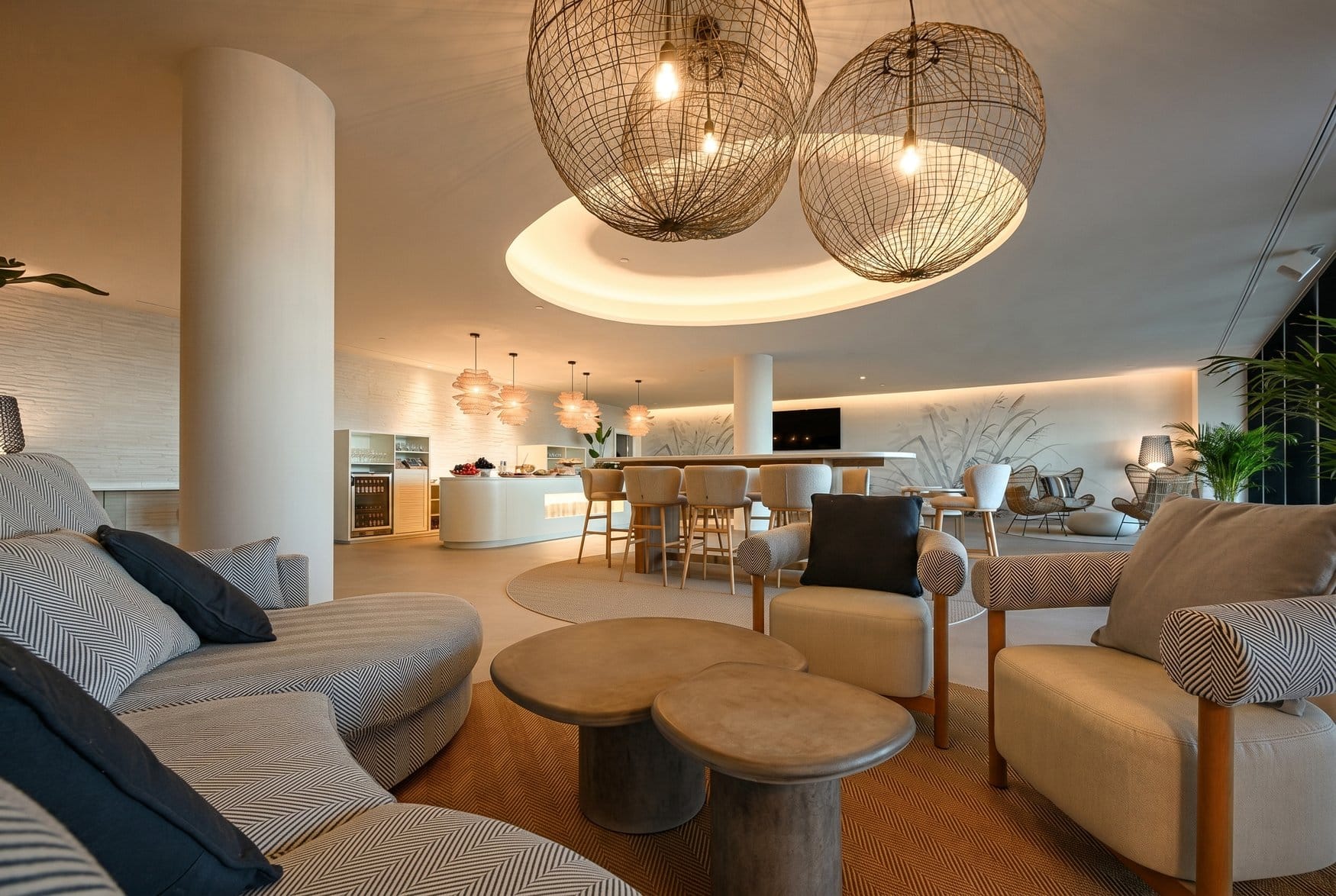 Die Lobby Lounge des Amáre Beach Hotel Sancti Petri mit drei großen Kugelleuchten aus Geflecht über einer runden Deckenmulde. Vorn stehen Polstersessel mit Fischgrätmuster um niedrige Steintische, dahinter eine Bar mit Hockern und ein Buffet.