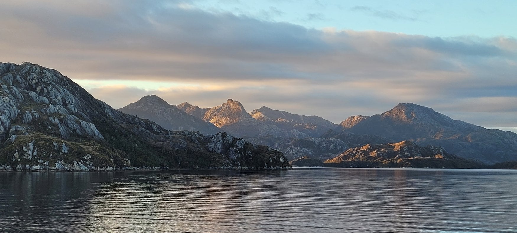 Die Magellanstraße im Süden Chiles im Abendlicht. Kahle Granitberge säumen das spiegelglatte Wasser, die tiefstehende Sonne färbt die Gipfel warm.