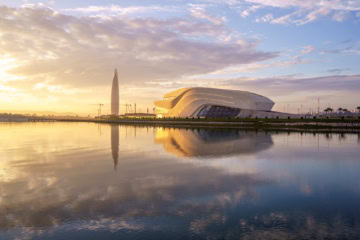 Das Grand Théâtre in Rabat am Ufer des Bouregreg in der Abenddämmerung. Der geschwungene weiße Baukörper liegt hinter Rasenflächen und einem Parkplatz, dahinter das Stadtbild von Salé.