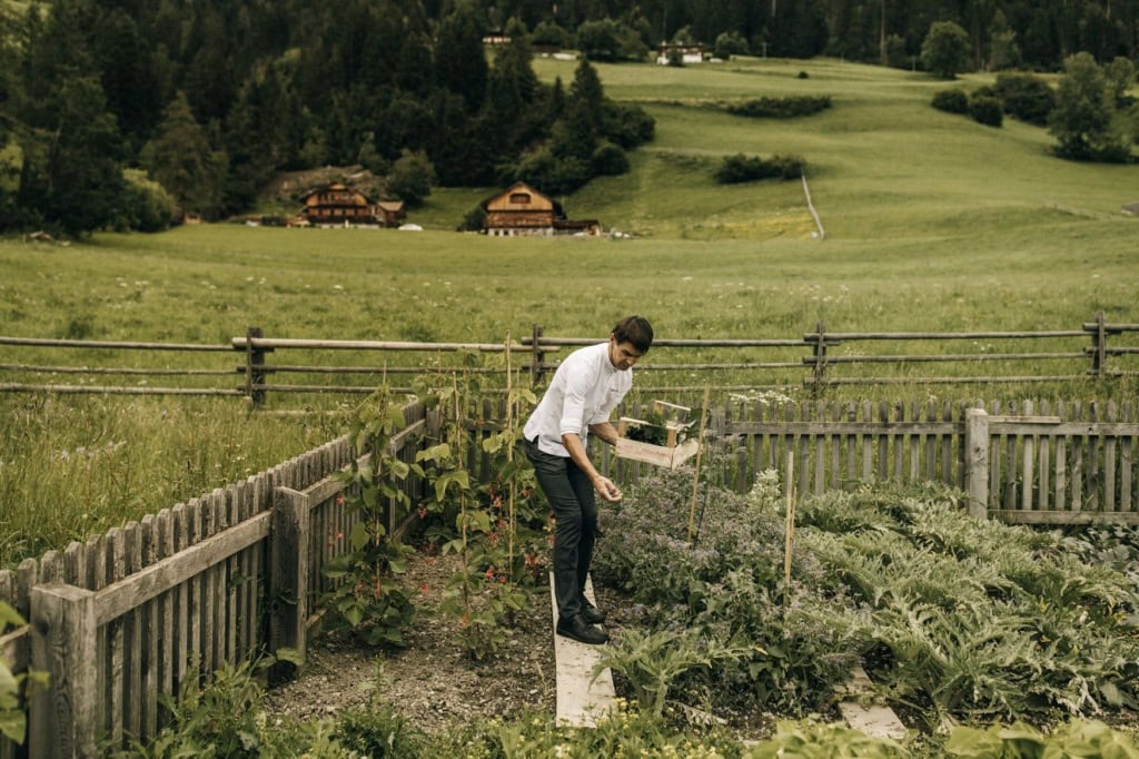 Küchenchef Markus Auer geht in weißer Kochjacke über einen Holzsteg im eingezäunten Gemüsegarten des Naturhotel Leitlhof und erntet Kräuter in eine Holzkiste. Dahinter liegen Bergwiesen mit Höfen.