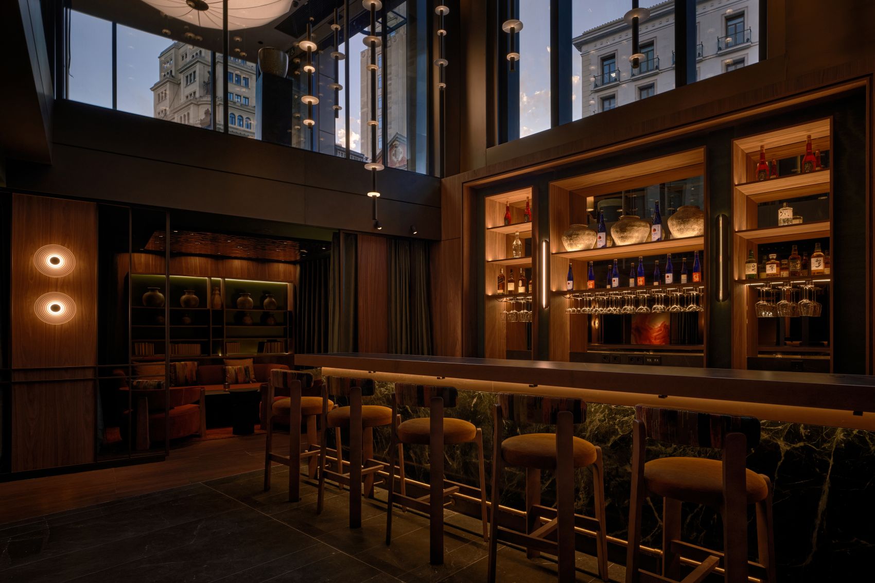 Bar im Nobu Hotel Madrid mit dunklen Holzelementen, beleuchteten Regalen und hohen Fenstern zur Madrider Innenstadt.