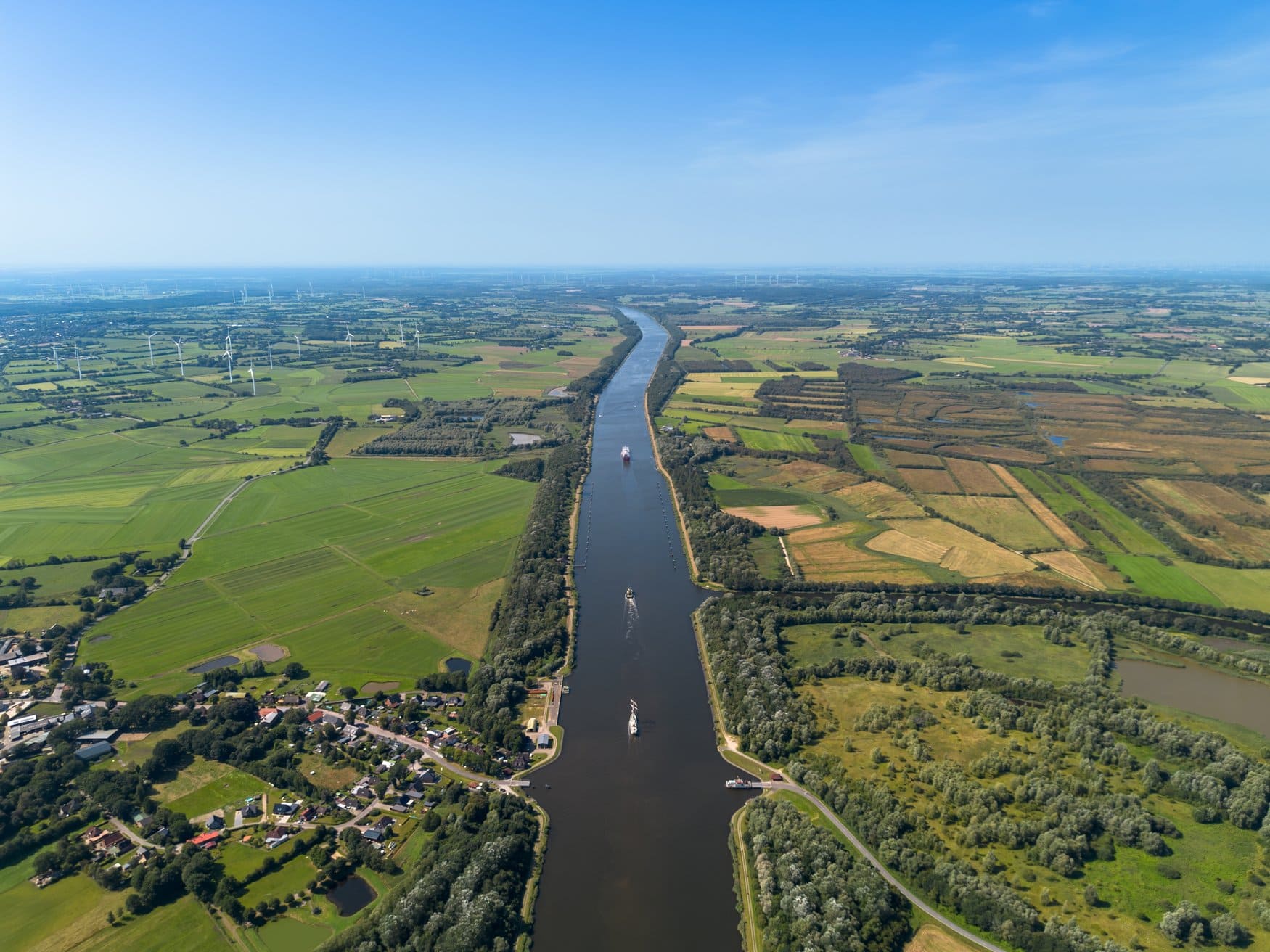 Luftaufnahme des Nord-Ostsee-Kanals in Schleswig-Holstein. Die gerade Wasserstraße zieht sich durch Felder und Weiden bis zum Horizont, auf ihr fahren ein Frachter und kleinere Boote.