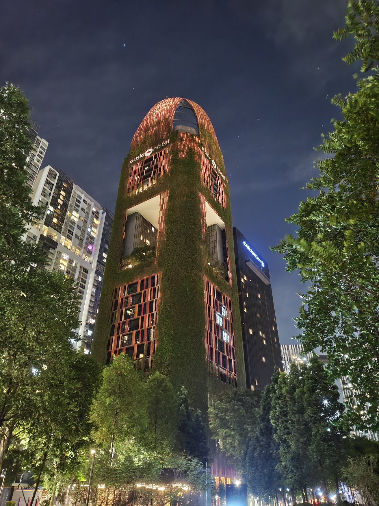 Der Turm des Oasia Hotel Downtown bei Nacht. Die rot-orangefarbene Gitterfassade ist dicht mit Kletterpflanzen bewachsen, links und rechts rahmen Bäume das Bild.