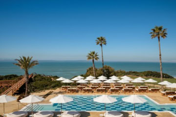 Blick über einen Pool mit blau-weißem Schachbrettmuster auf dem Boden. Dahinter stehen Reihen von Holzliegen unter weißen Sonnenschirmen, links führt eine Holztreppe zum Strand, rechts liegt ein Golfgrün. Über Palmen hinweg geht der Blick aufs Meer.