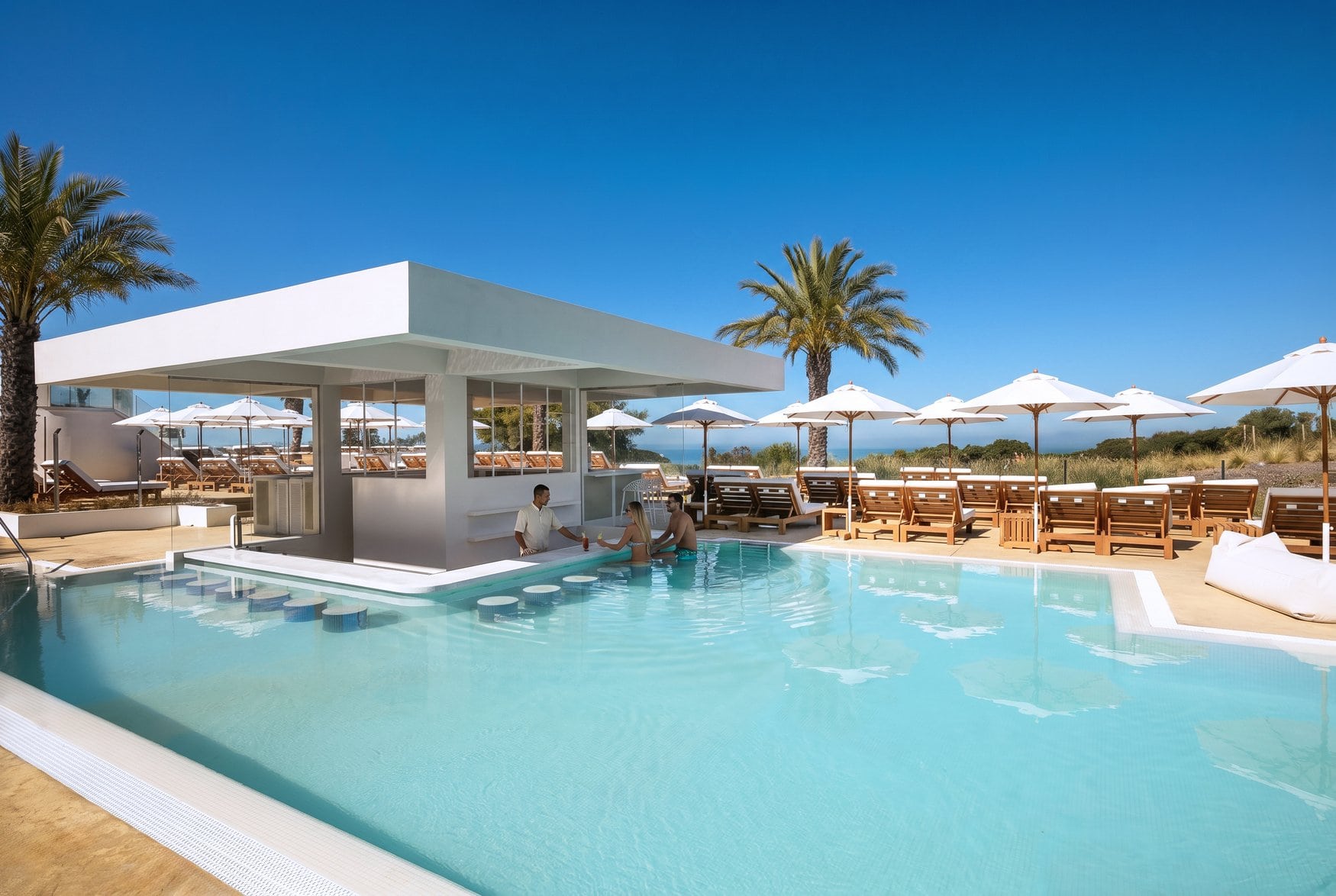 Eine Poolbar mit flachem weißem Dach steht im Wasser, davor sitzen zwei Gäste auf Barhockern im Pool und werden vom Barkeeper bedient. Rechts reihen sich Holzliegen unter weißen Sonnenschirmen.