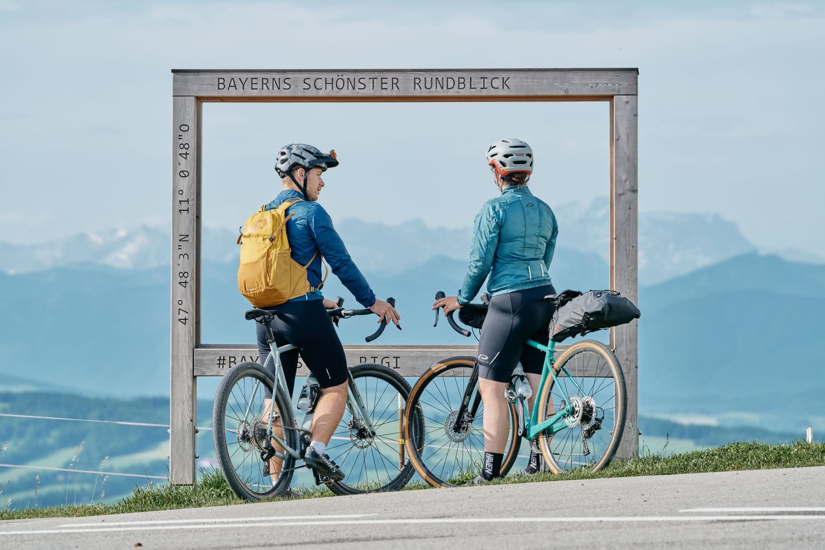 Zwei Gravelbiker genießen vom Hohen Peißenberg den Blick auf die bayerischen Alpen.