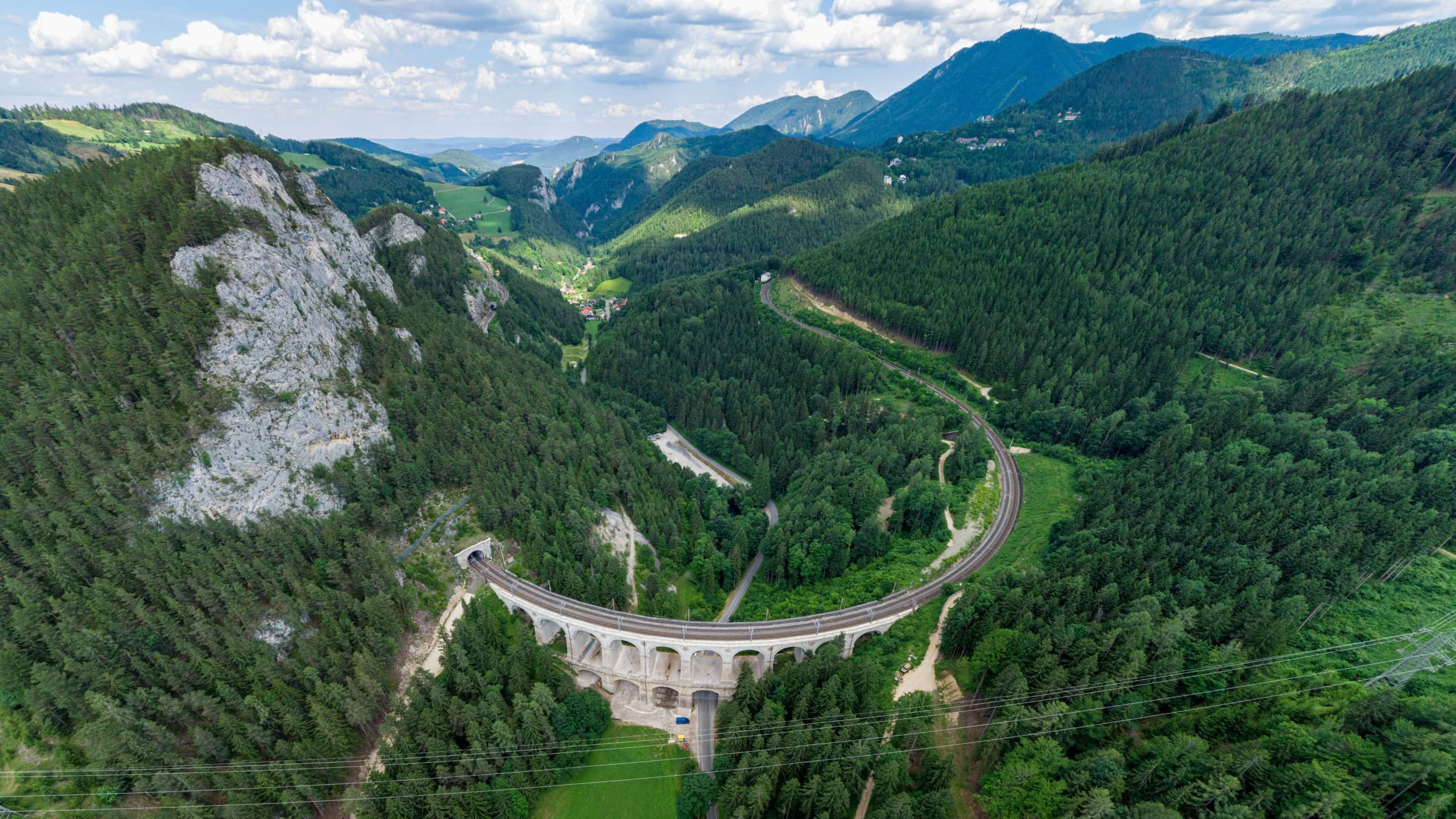 Luftaufnahme eines Viadukts der Semmeringbahn, das sich in einer Schleife durch bewaldetes Bergland zieht, mit Tunnelportal am Fuß des Felsens.