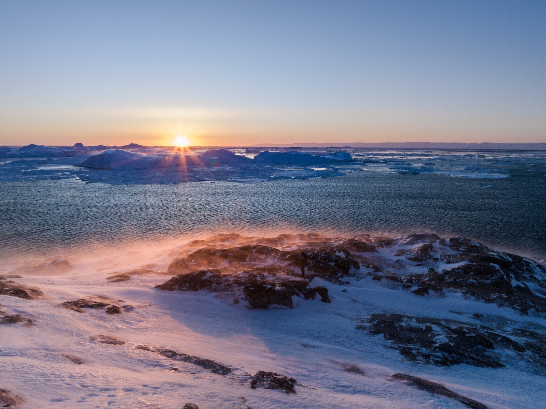 Luftaufnahme bei Sonnenaufgang über dem Eisfjord. Die tiefstehende Sonne färbt Schnee und Felsen orange, auf dem Wasser liegen Eisberge.