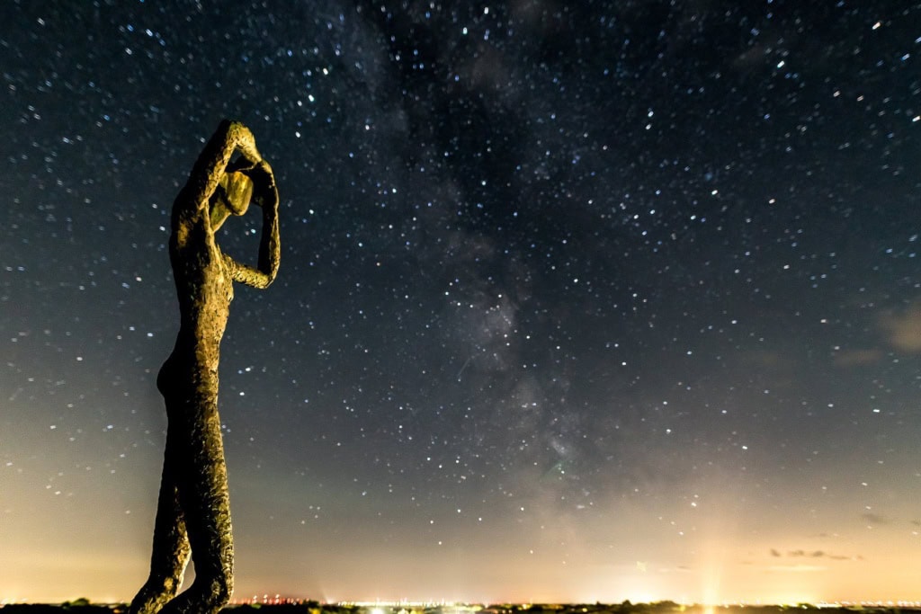 Die angestrahlte Skulptur Utkieker auf Spiekeroog steht vor dem Nachthimmel, hinter ihr zieht sich das Band der Milchstraße. Am unteren Bildrand leuchten Ortslichter. 