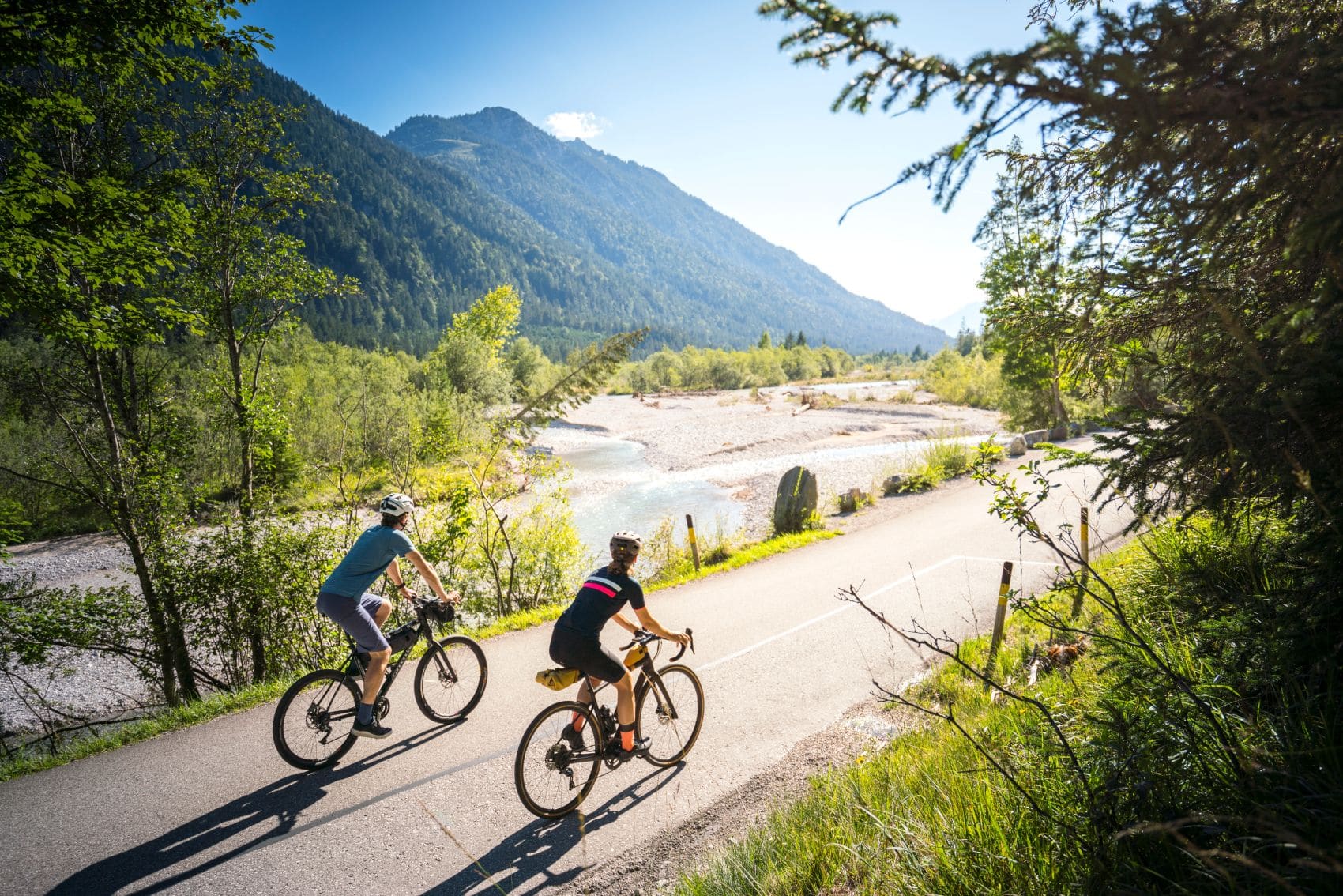 Zwei Radfahrer fahren auf einem Wasser-Radlweg durch die Berglandschaft Oberbayerns.