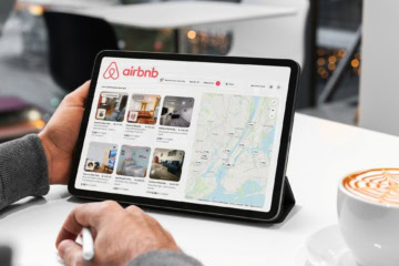 Eine Person hält in einem Café ein Tablet mit der Airbnb-Unterkunftssuche für New York. Auf dem Bildschirm sind sechs Angebote mit Preisen und Bewertungen sowie eine Karte mit Preismarkierungen zu sehen, rechts steht eine Tasse Kaffee.