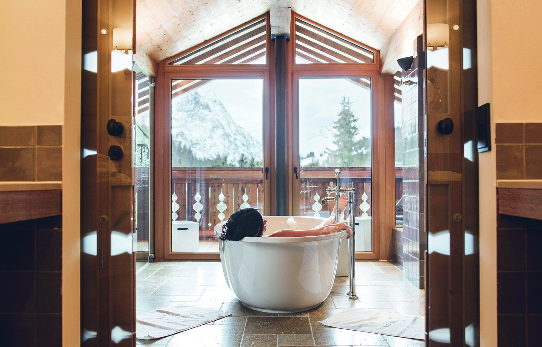 Blick durch eine geöffnete Flügeltür in ein Badezimmer mit Natursteinboden. In der freistehenden Wanne liegt eine Person mit dem Rücken zur Kamera, durch die Giebelfenster dahinter ist ein schneebedeckter Berg zu sehen.