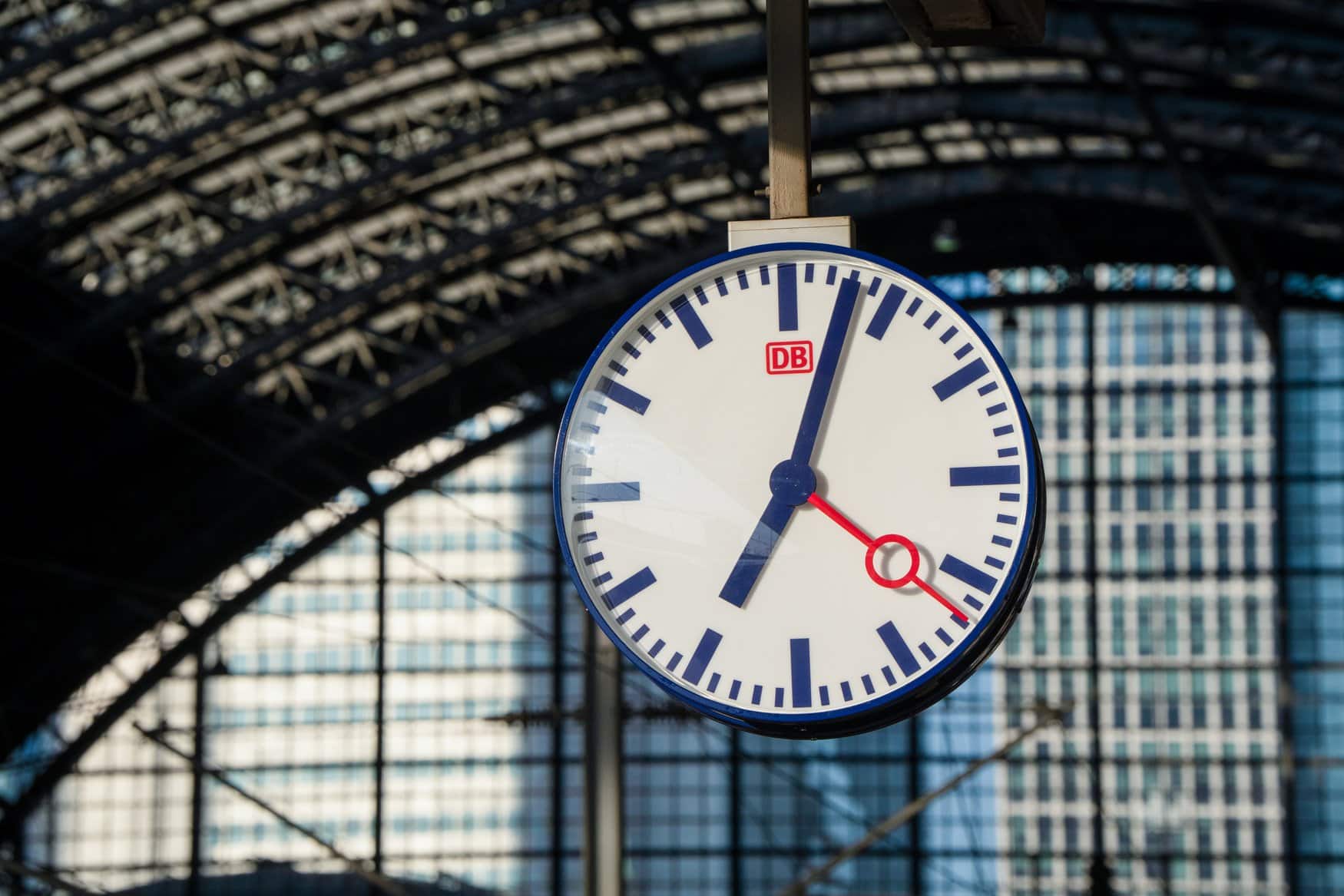 Eine Bahnhofsuhr mit blauem Rahmen, dunkelblauen Zeigern und rotem Sekundenzeiger hängt unter der Stahlkonstruktion der Bahnsteighalle im Frankfurter Hauptbahnhof. Auf dem Zifferblatt sitzt das rote DB-Logo.