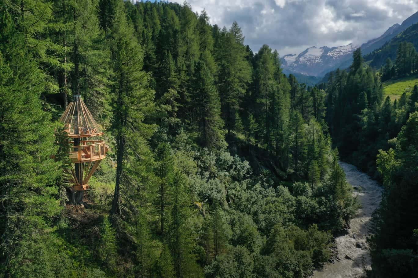 Das Baumhaus Ochsenkopf zwischen hohen Lärchen und Zirben am Hang über einem Gebirgsbach, im Hintergrund schneebedeckte Gipfel der Ötztaler Alpen.