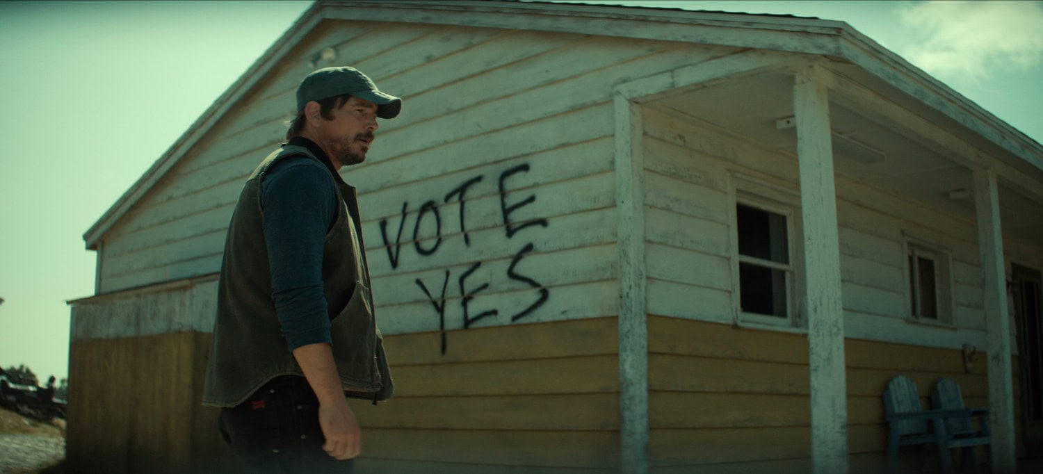 Szene aus »Below«: Ein Mann mit Kappe geht an einem Holzhaus vorbei, auf dessen Wand »Vote Yes« gesprüht steht, ein Verweis auf die Abstimmung über die Umsiedlung.