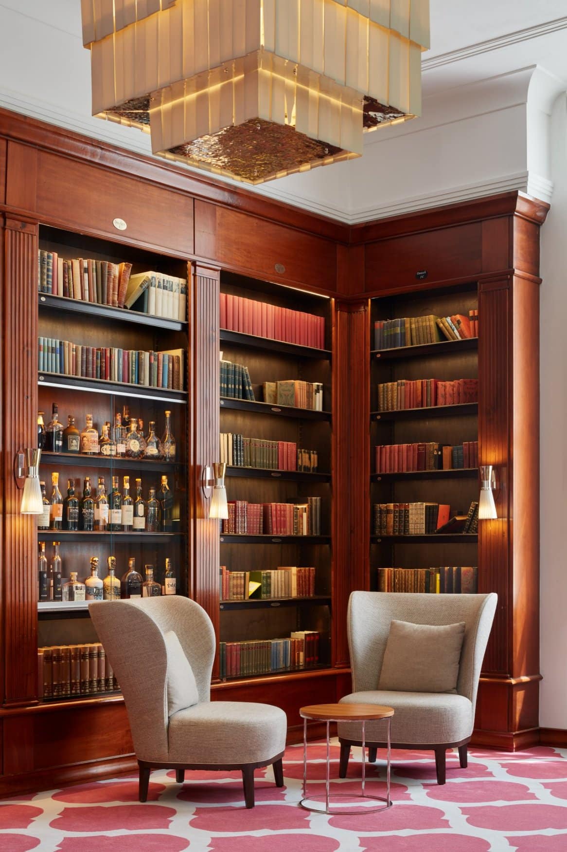 Bibliothek mit deckenhohen Bücherregalen aus Kirschholz zwischen hohen Fenstern mit Vorhängen. Auf einem pink gemusterten Teppich stehen Polstersessel und Beistelltische, links ein Lesetisch mit Blumenstrauß und aufgeschlagenen Bildbänden. Über der Sitzgruppe hängt ein eckiger Leuchter.