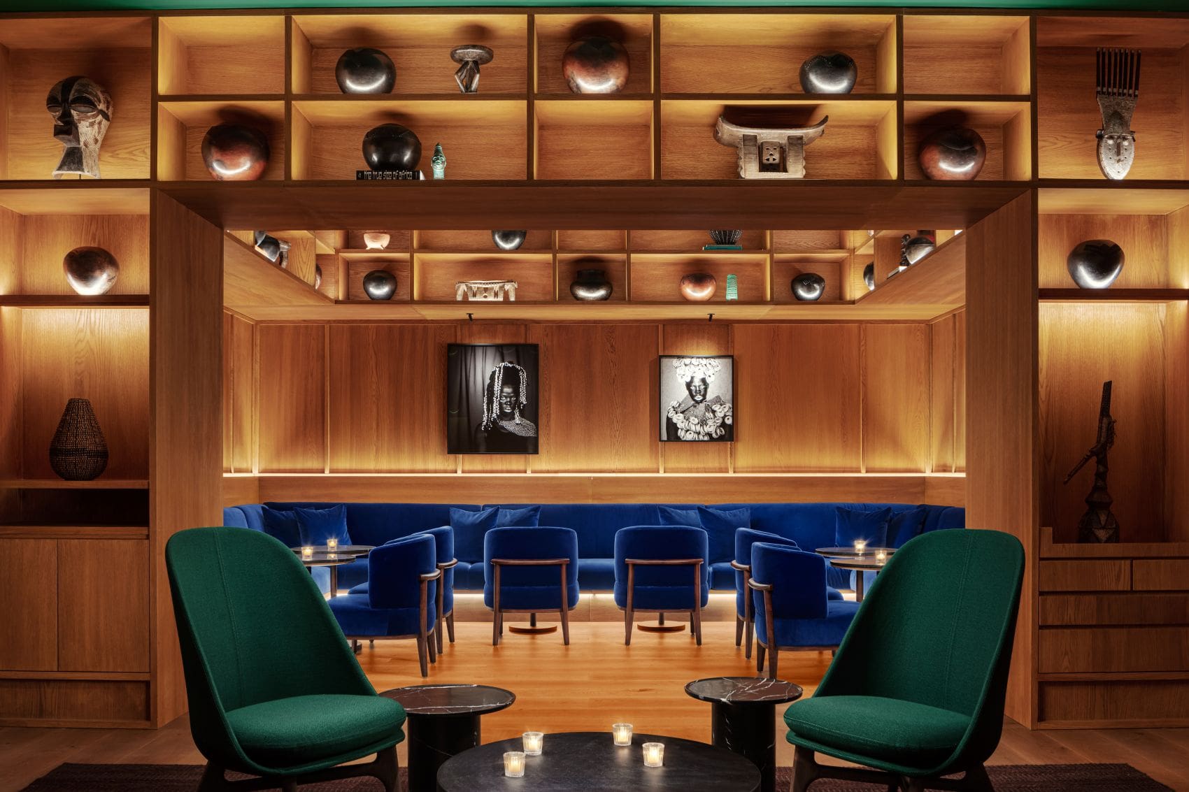 Punch Room im Cape Town EDITION mit blauen Samtsofas, grünen Lounge-Sesseln, beleuchteten Holzregalen und afrikanischen Kunstwerken.