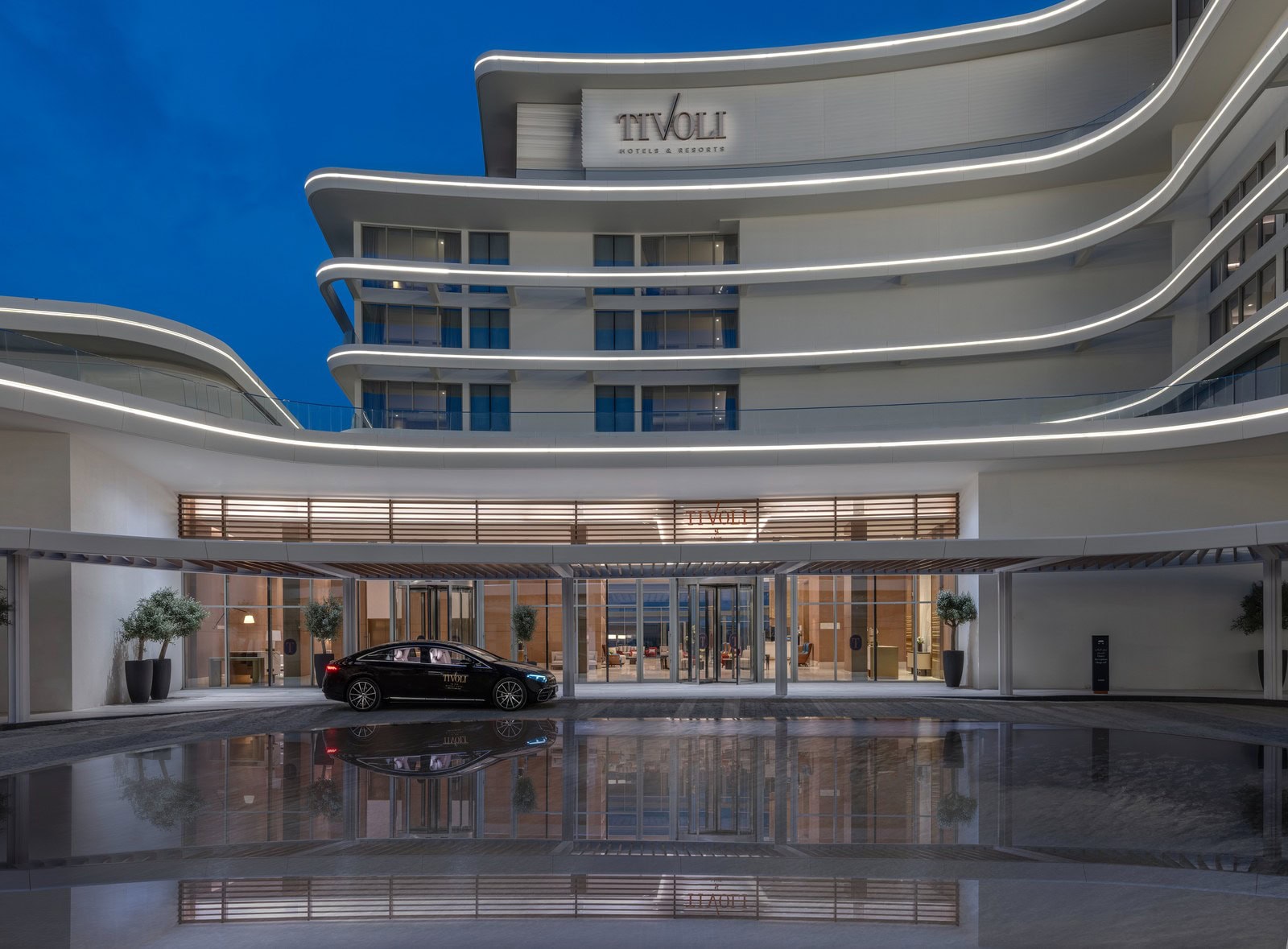 Geschwungene weiße Fassade des Tivoli LA VIE Muscat mit beleuchteten Lichtbändern, davor die Vorfahrt mit Wasserbecken in der Abenddämmerung