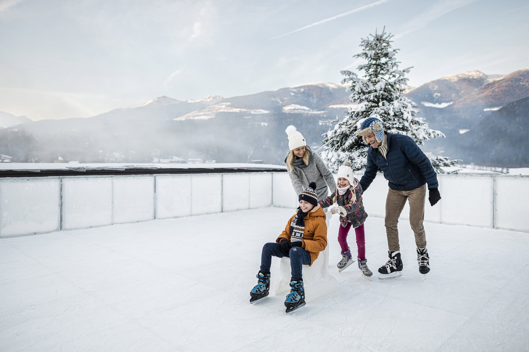 Familie beim Eislaufen auf dem Dach des Falkensteiner Family Resort Lido vor verschneiter Bergkulisse.