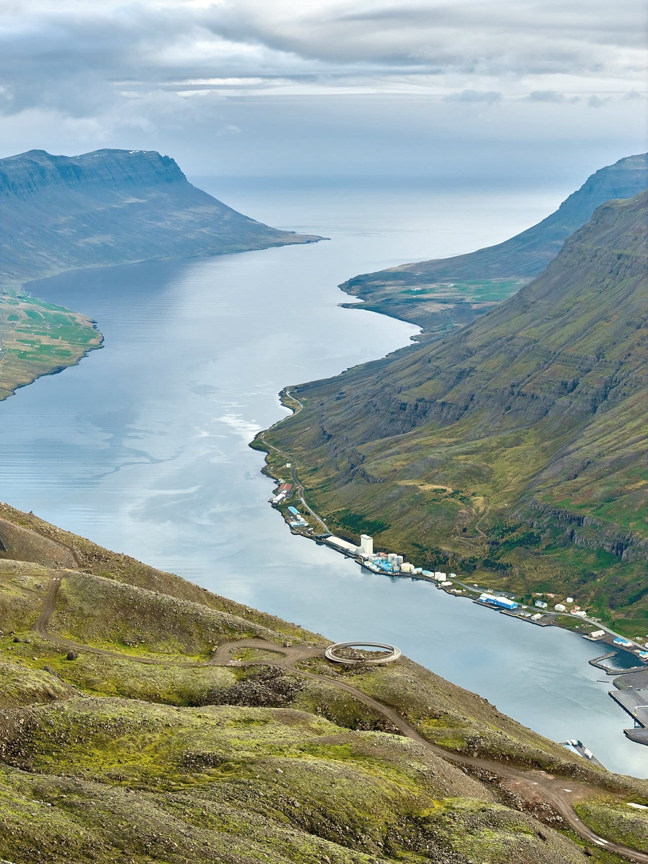 Blick von einem Berghang über einen Fjord. Am rechten Ufer liegt eine Ortschaft mit Hafenanlagen, Pfade ziehen sich über die Kuppe im Vordergrund, am Horizont öffnet sich das Meer.