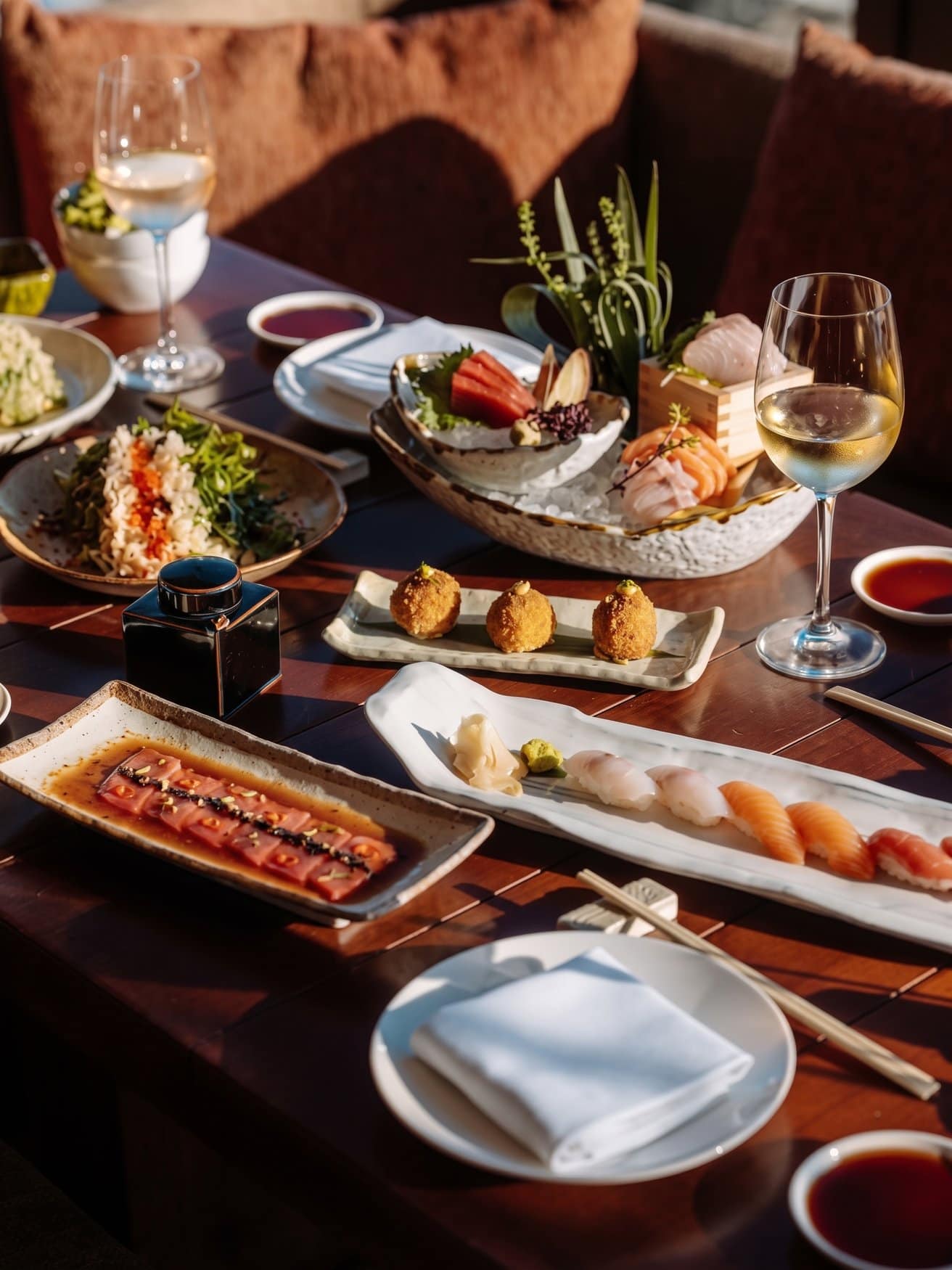 Ein dunkler Holztisch im Abendlicht mit mehreren japanischen Gerichten: Sashimi auf Eis in einer Muschelschale, eine Reihe Nigiri auf einer langen Platte, mariniertes Thunfisch-Carpaccio, frittierte Bällchen und zwei Gläser Weißwein.