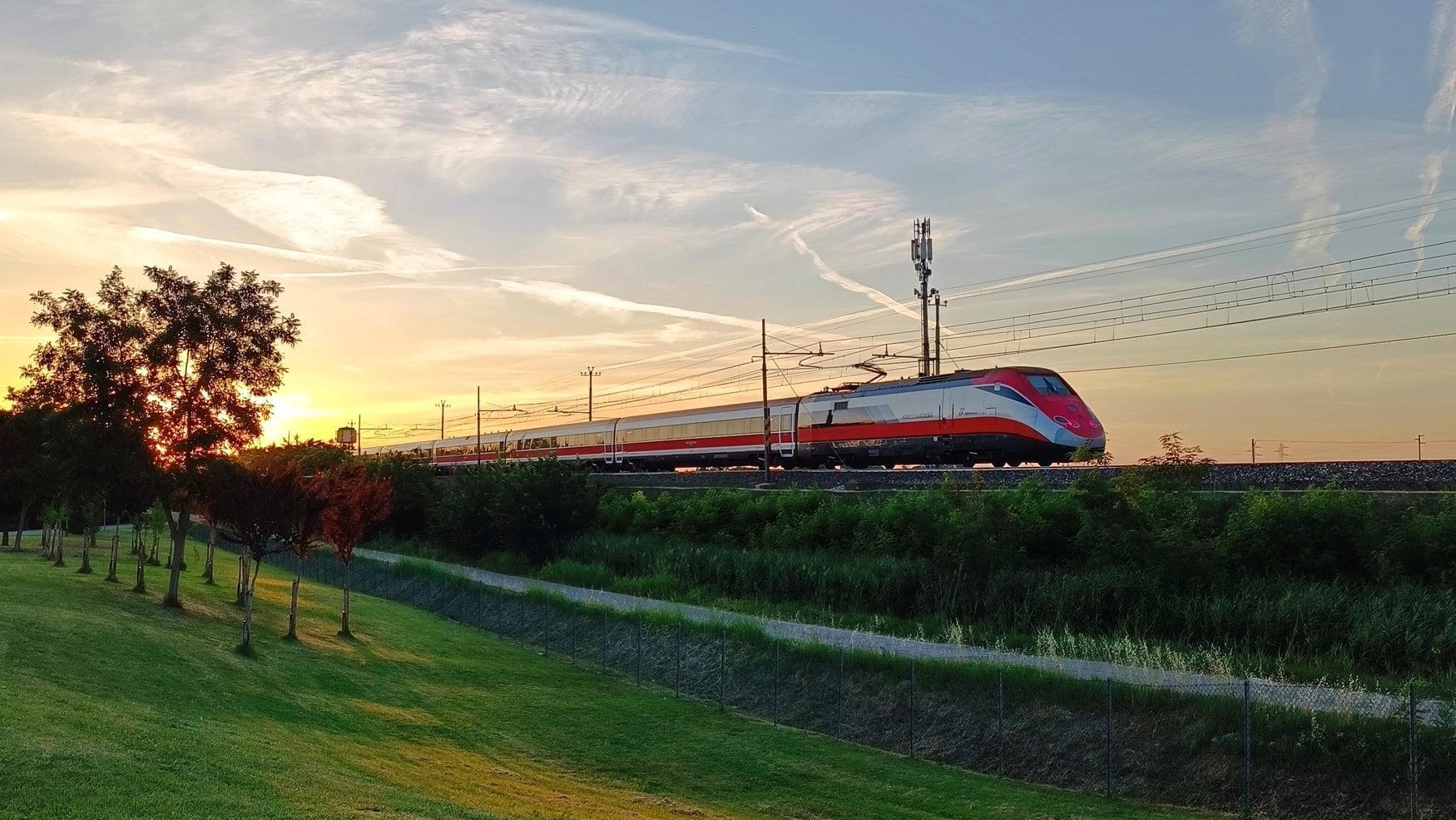 Ein Frecciarossa der Trenitalia fährt im Sonnenuntergang auf einer Strecke durch flaches Land bei Faenza. Im Vordergrund liegen eine Wiese mit jungen Bäumen und ein Zaun entlang der Gleise.