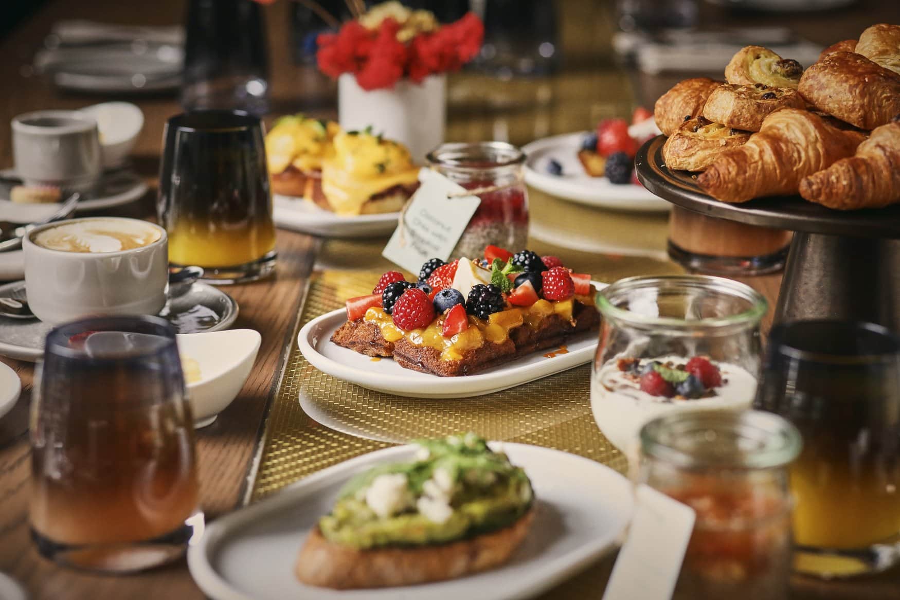 Gedeckter Frühstückstisch mit einer Schnitte voll Beeren und Mangocreme in der Mitte, davor Avocadobrot mit Feta. Rechts stapeln sich Croissants und Plundergebäck auf einer Etagere, dazwischen Kaffee, Saft und Joghurt im Glas.