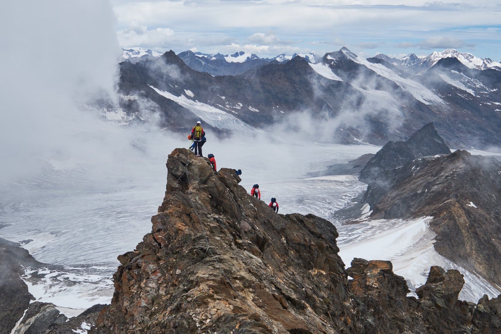 Eine Gruppe von Bergsteigern mit Helm klettert über einen felsigen Grat, unter ihnen breiten sich Gletscher und Wolkenfelder aus.