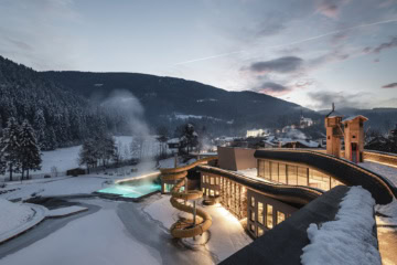 Falkensteiner Family Resort Lido im Winter mit beleuchtetem Außenpool, Wasserrutsche und verschneiter Berglandschaft.