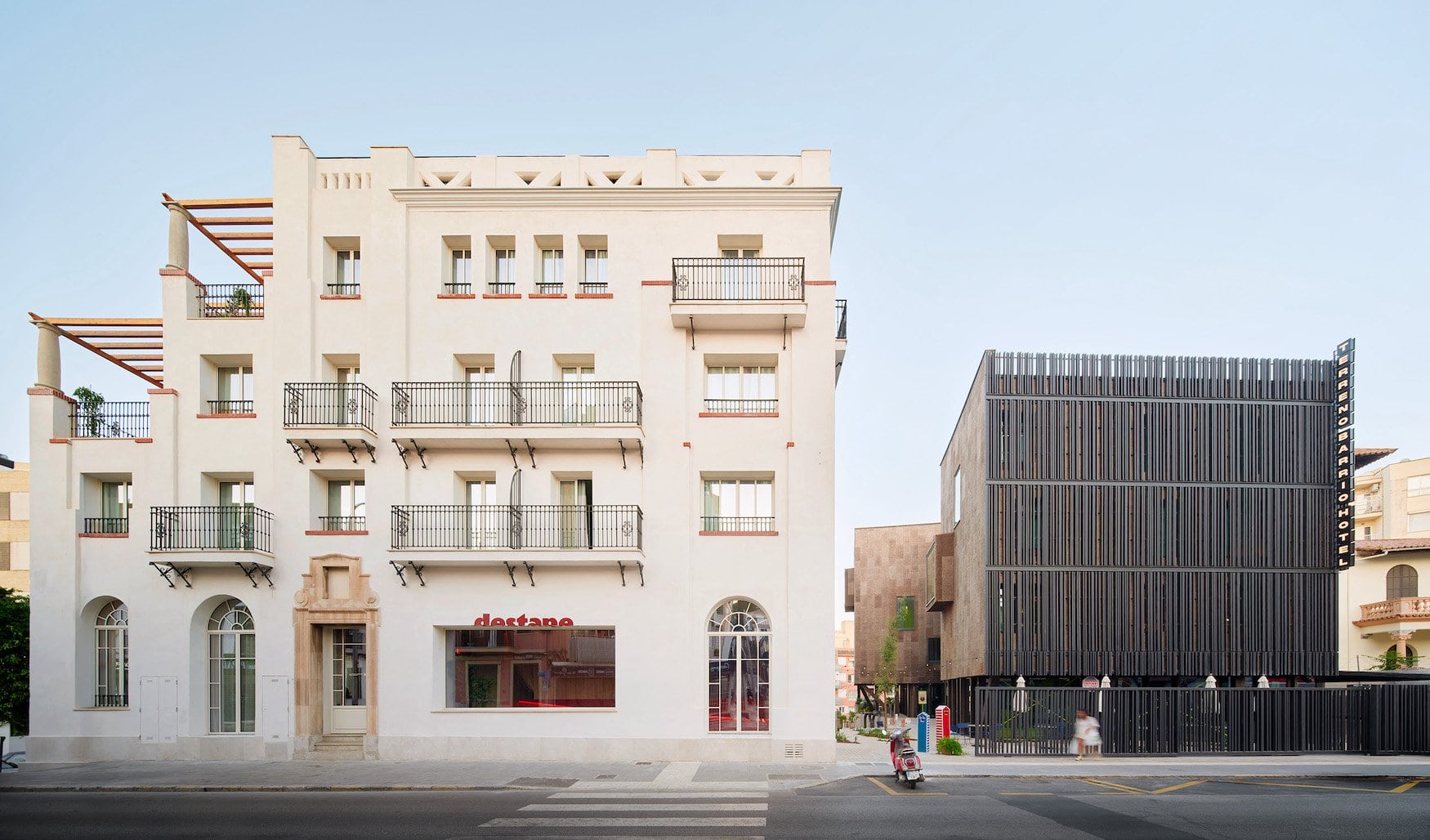 Straßenansicht eines weiß verputzten Altbaus mit schmiedeeisernen Balkonen und Dachpergolen, im Erdgeschoss der rote Schriftzug „destape”. Rechts daneben steht ein dunkler Neubau mit vertikaler Lattenfassade und dem Schriftzug „Terreno Barrio Hotel”.