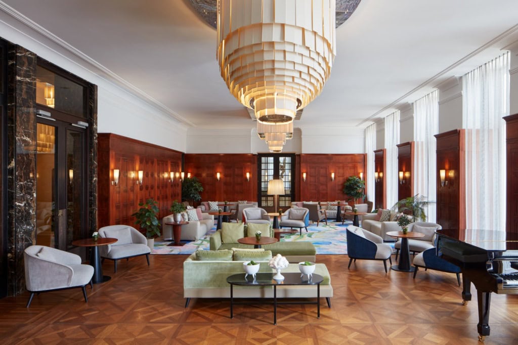 Hohe Hotellobby mit holzvertäfelten Wänden und Wandleuchten, darüber hängt ein großer gestufter Kronleuchter. Auf Fischgrät- und Tafelparkett stehen Sitzgruppen aus hellen Polstersesseln und grünen Samtsofas um runde Tische, rechts ein schwarzer Flügel.