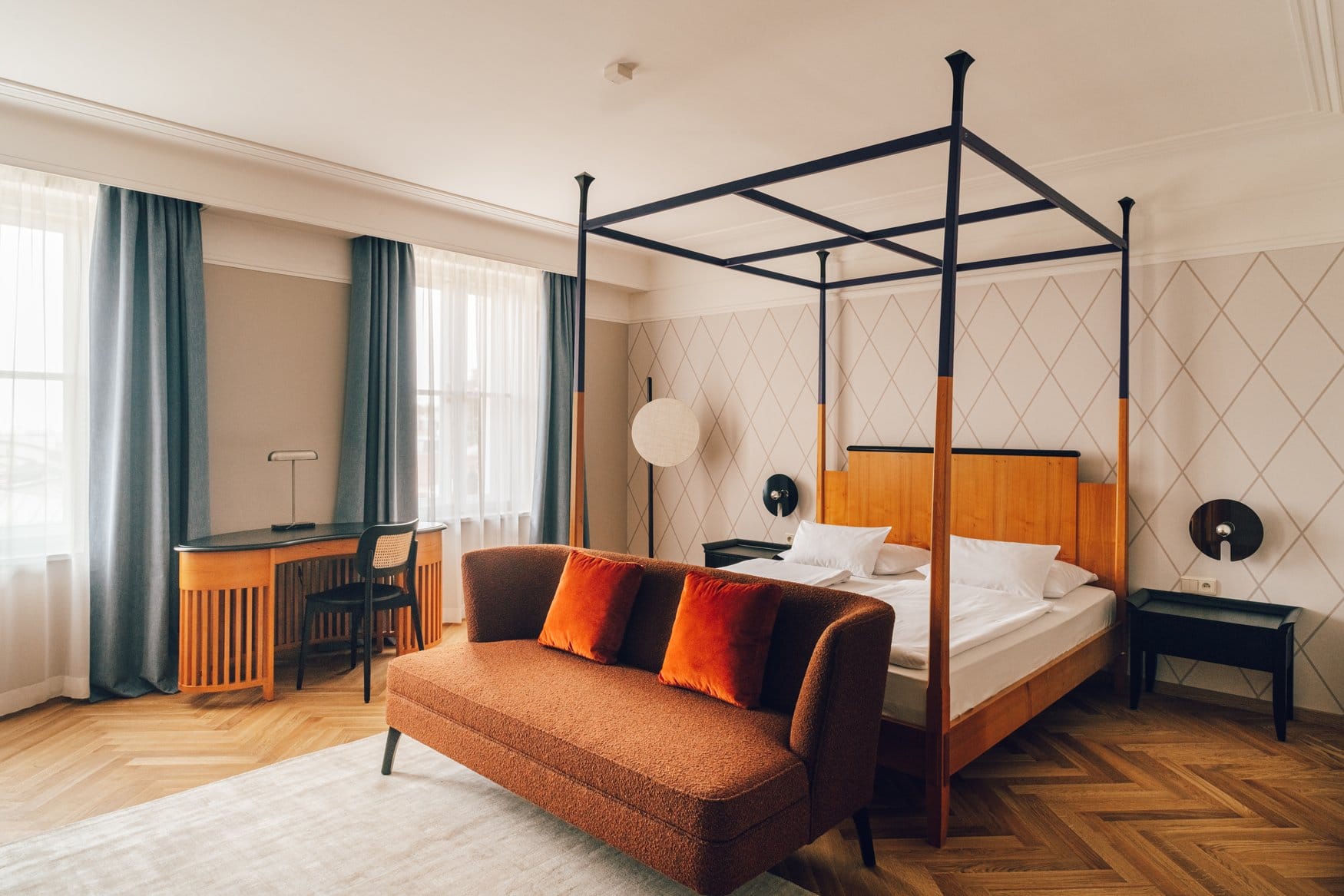 Hotelzimmer mit Himmelbett aus Holz und schwarzem Metallrahmen vor einer Wand mit Rautenmuster. Davor steht ein rostrotes Sofa mit orangefarbenen Kissen, links am Fenster ein geschwungener Schreibtisch mit Stuhl, auf dem Boden Fischgrätparkett.