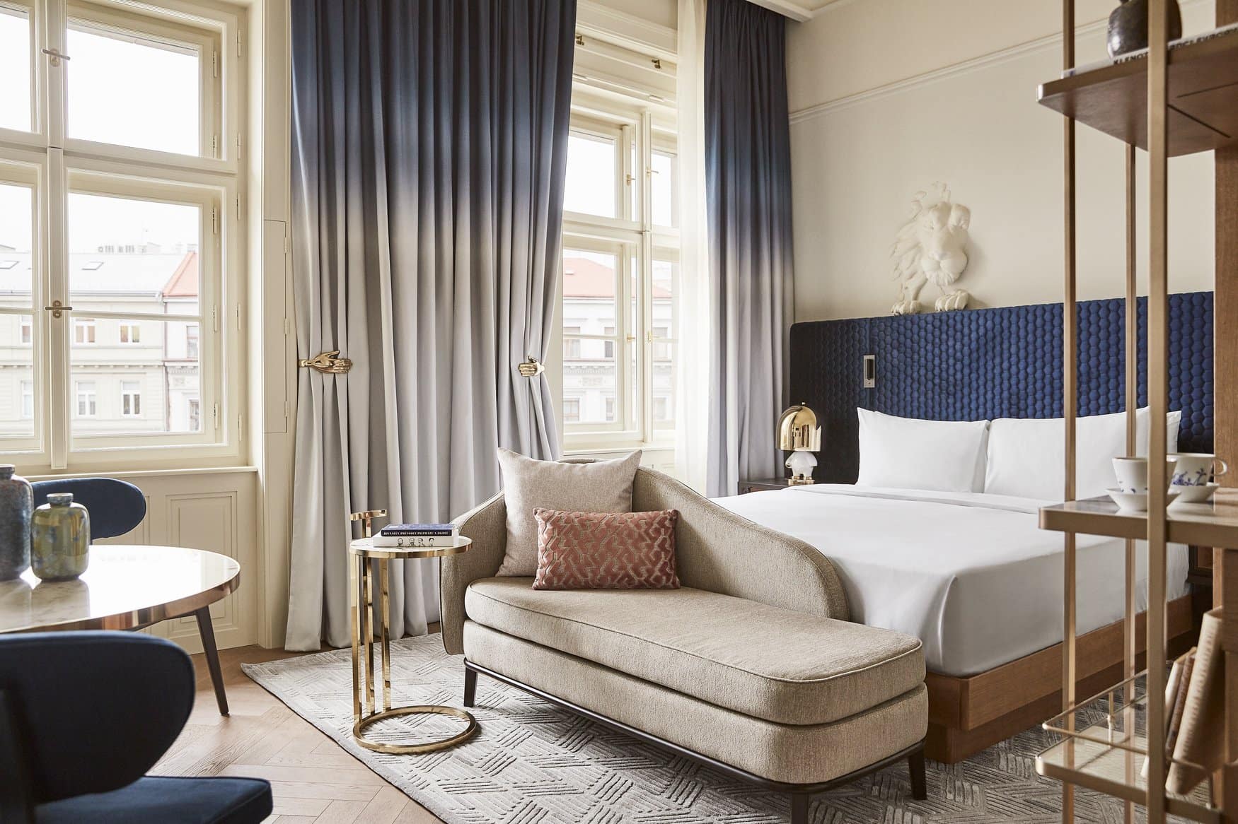 Hotelzimmer mit Doppelbett vor blauem Kopfteil, darüber eine weiße Löwenfigur. Davor steht eine beige Récamiere, links fällt Licht durch hohe Fenster mit Vorhängen im Farbverlauf.