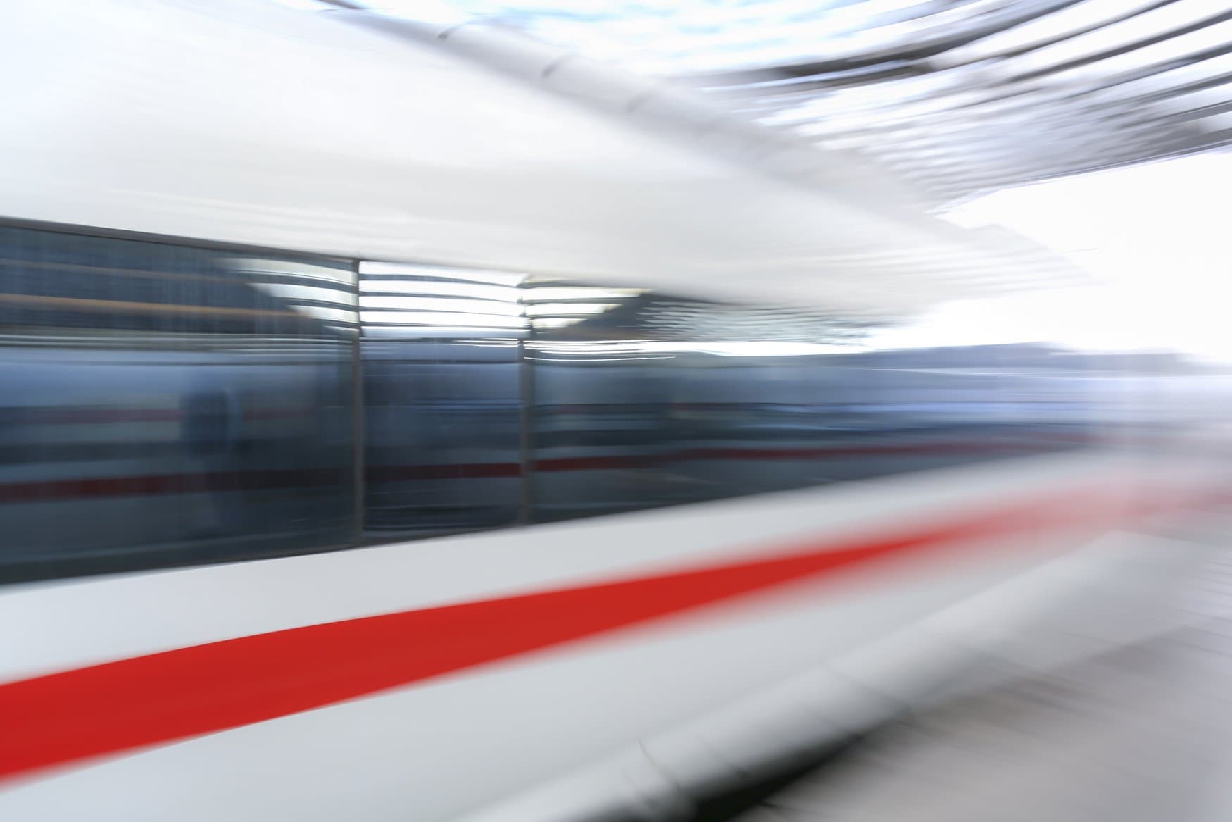Ein ICE fährt mit hohem Tempo durch einen Bahnhof, Wagenkasten und Hallendach verwischen zu horizontalen Streifen. Der rote Zierstreifen zieht sich als Linie durch das Bild.