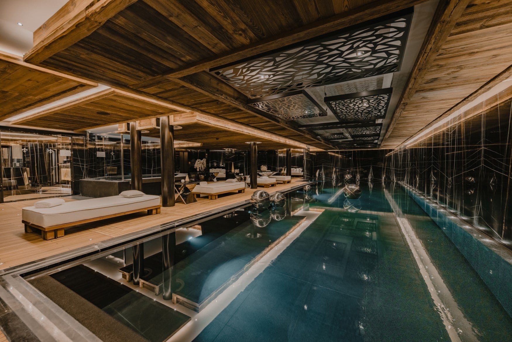 Ein länglicher Innenpool unter einer Decke aus Altholzbalken mit schwarzen Zierpaneelen. Links reihen sich weiße Liegen auf einem Holzdeck, die Wände sind mit schwarzem Marmor verkleidet und spiegeln sich im Wasser.