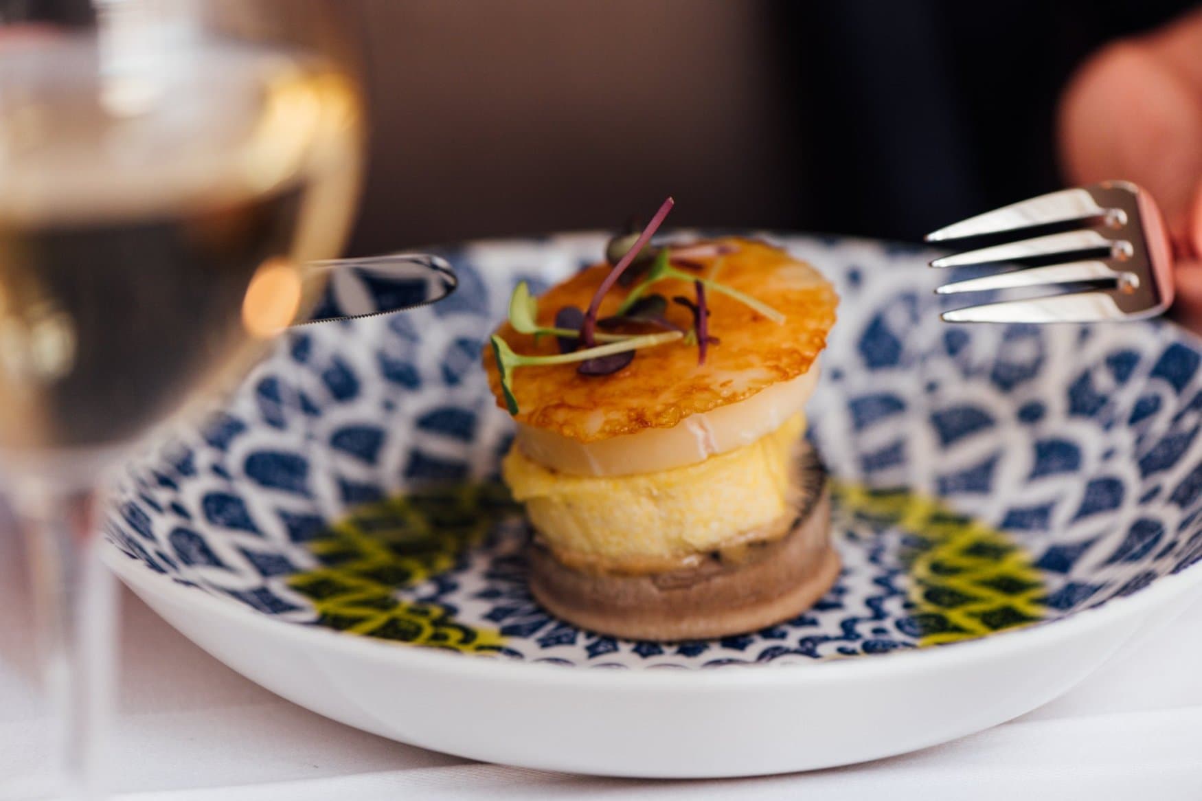Ein Türmchen aus Pilz, Creme und gratinierter Jakobsmuschel auf blau-grün gemustertem Teller, garniert mit Microgreens. Links steht ein Glas Weißwein, rechts hält eine Hand eine Gabel.