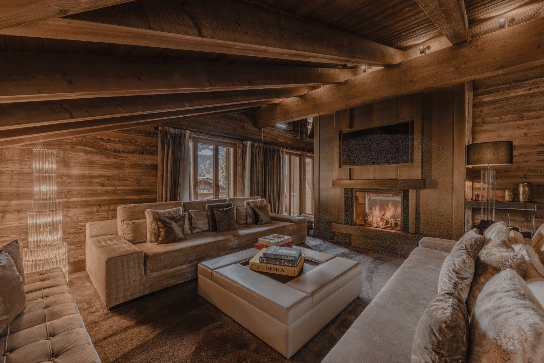 Ein Kaminraum mit Altholzwänden, in dem ein Feuer hinter einer Glasscheibe brennt, darüber ein Fernseher. Um einen quadratischen Lederhocker mit Bildbänden stehen Samtsofas mit Fell- und Samtkissen. Durch die Fenster ist das verschneite Tal zu sehen.