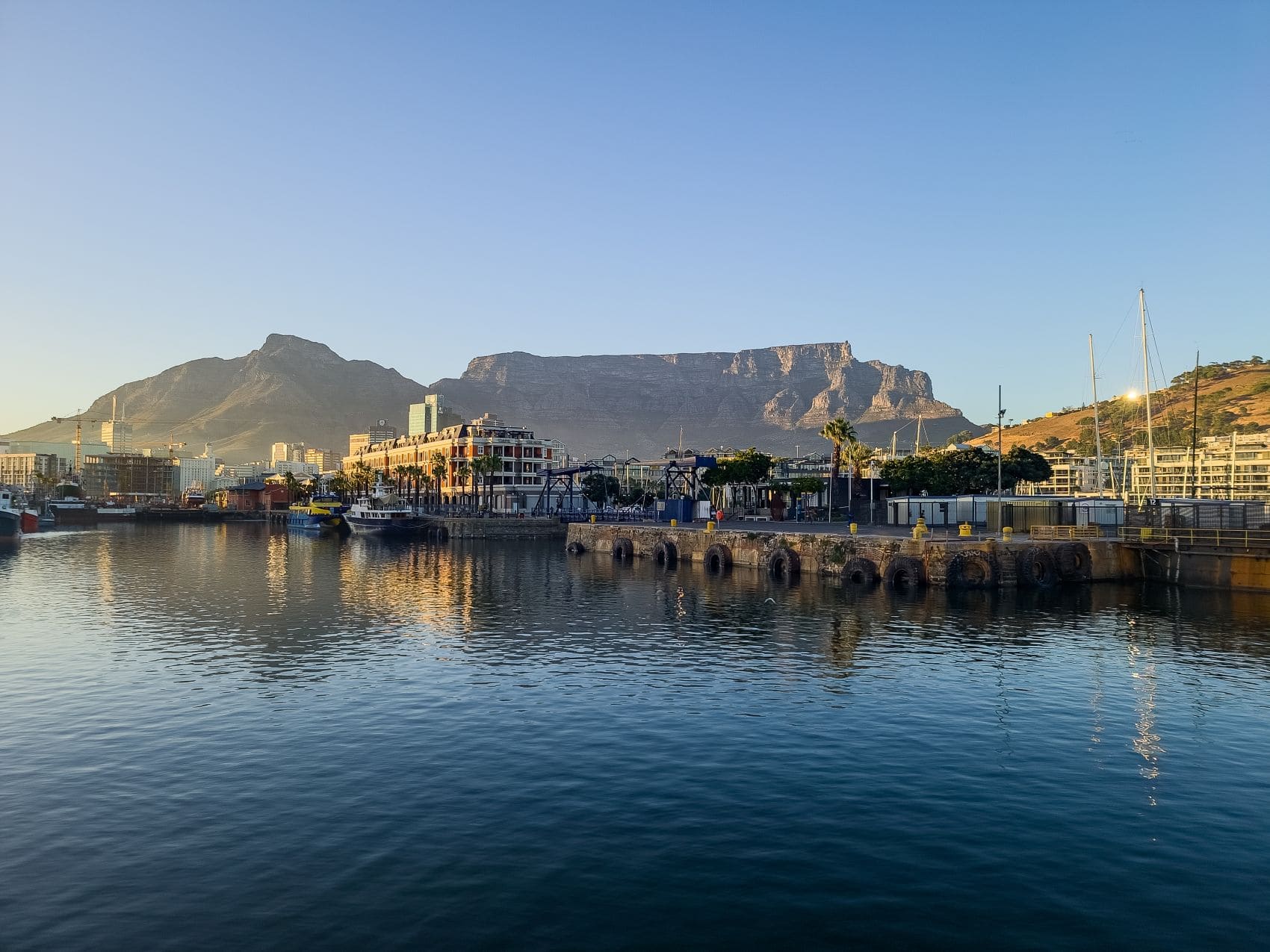 Blick über das Hafenbecken der V&A Waterfront in Kapstadt mit Hafenanlagen, Segelbootmasten und dem Tafelberg im Hintergrund.