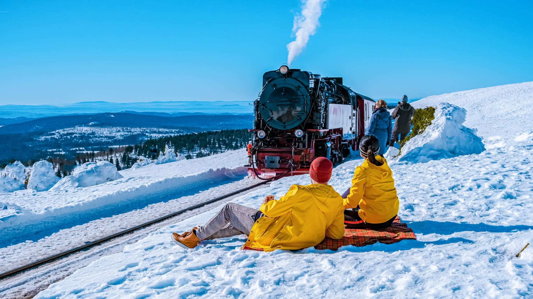 Ferien in Sachsen-Anhalt 2026: Dampflok der Brockenbahn auf verschneiter Strecke, davor zwei Personen in gelben Jacken im Schnee.