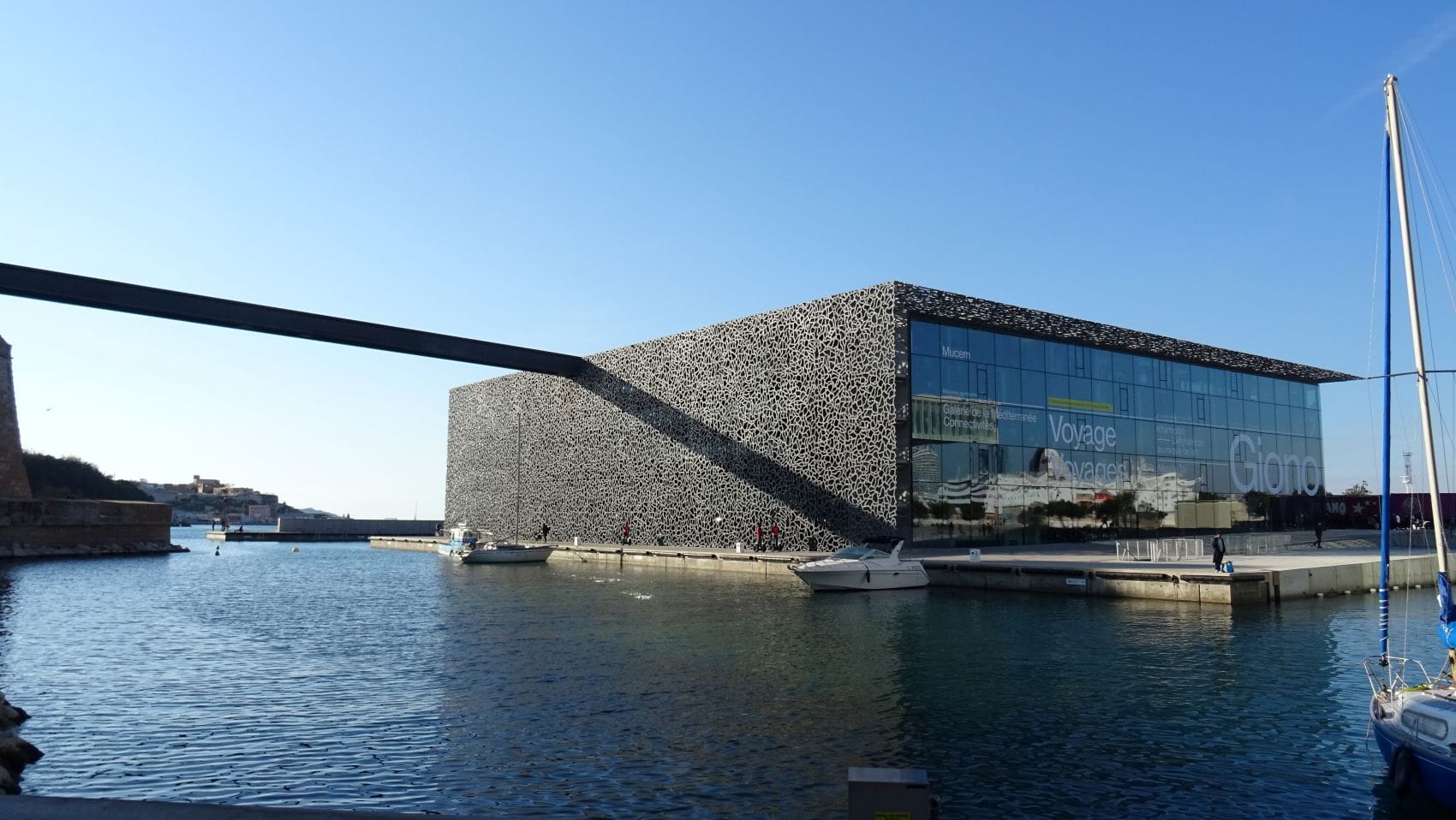 Mucem in Marseille mit seiner filigranen Betonfassade und großen Glasfront direkt am Wasser, im Vordergrund mehrere Segelboote.