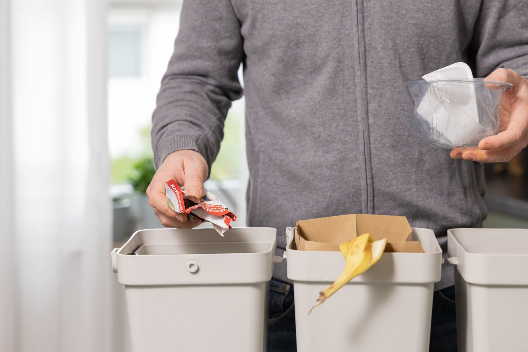 Eine Person in grauer Jacke wirft eine zerknüllte Verpackung in einen von drei hellen Abfallbehältern und hält in der anderen Hand eine Plastikschale. Im mittleren Behälter liegen ein Pappkarton und eine Bananenschale.