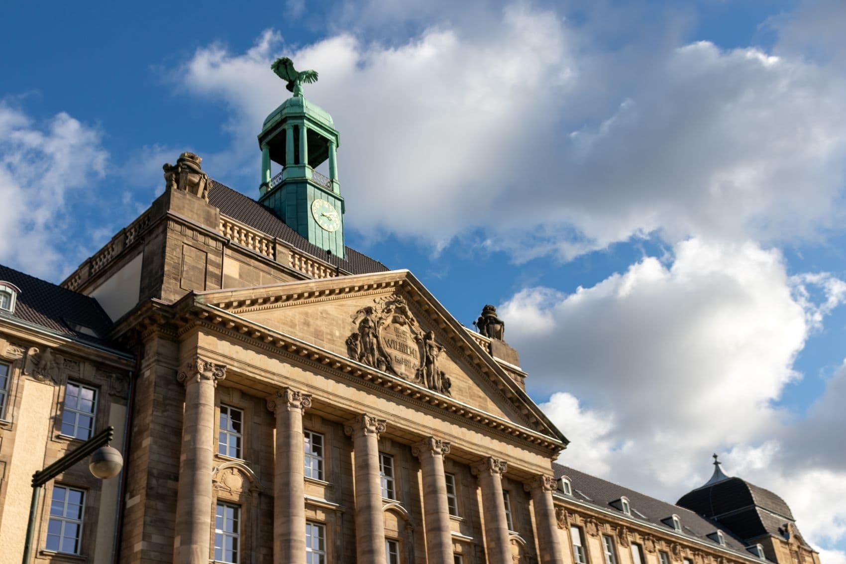 Historische Fassade des Oberlandesgerichts Düsseldorf mit Säulen, Giebel und grünem Uhrenturm vor blauem Himmel.