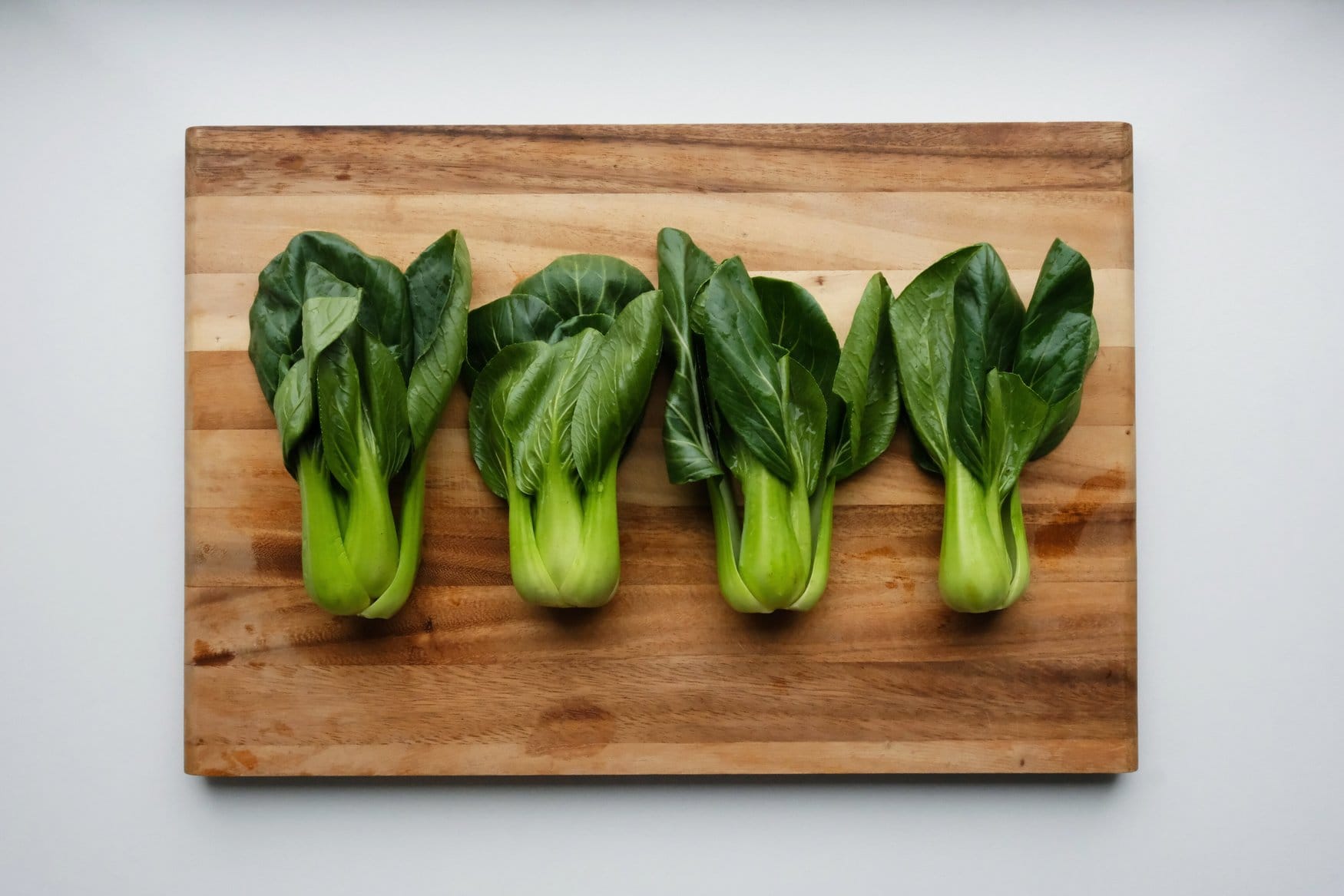 Vier Pak-Choi-Stauden mit hellgrünen Stielen und dunklen Blättern liegen nebeneinander auf einem Schneidebrett aus Holz.
