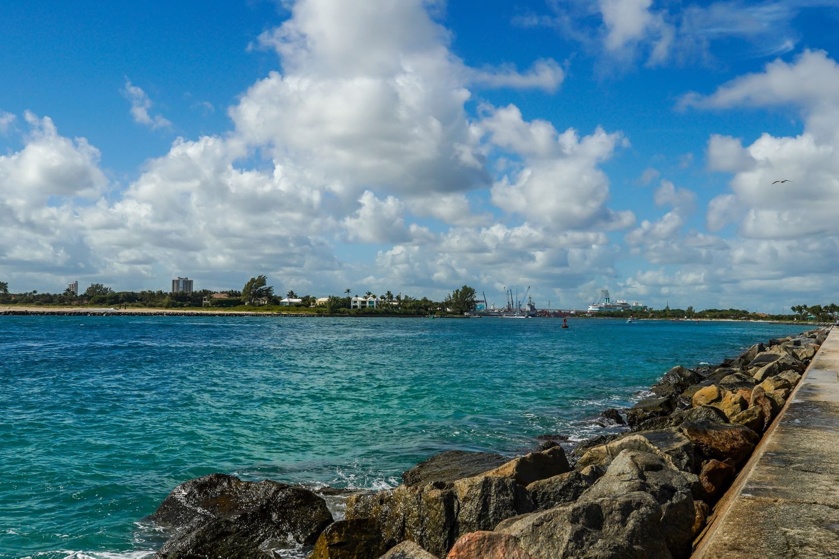 Blick über das türkisfarbene Wasser des Palm Beach Inlet in Florida mit felsiger Uferbefestigung, Schiffen in der Ferne und blauem Himmel.