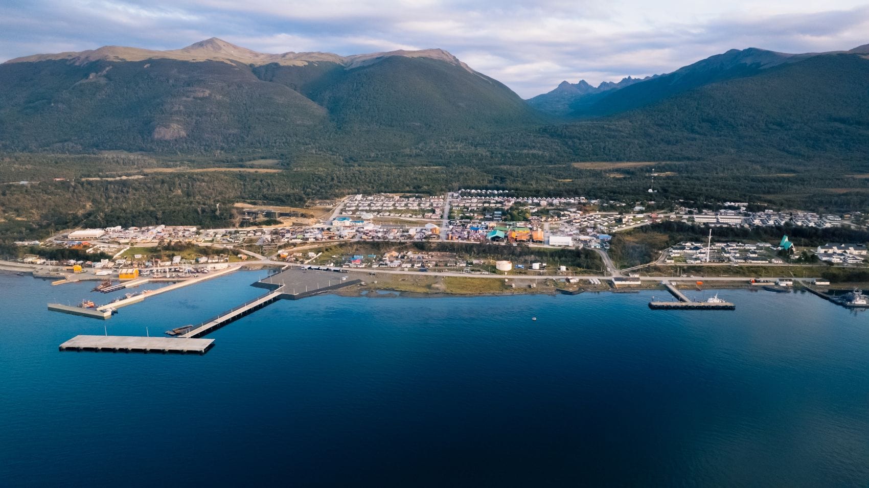 Luftaufnahme von Puerto Williams in Chile mit Hafenanlagen am Beagle-Kanal, Wohnhäusern und bewaldeten Bergen im Hintergrund.