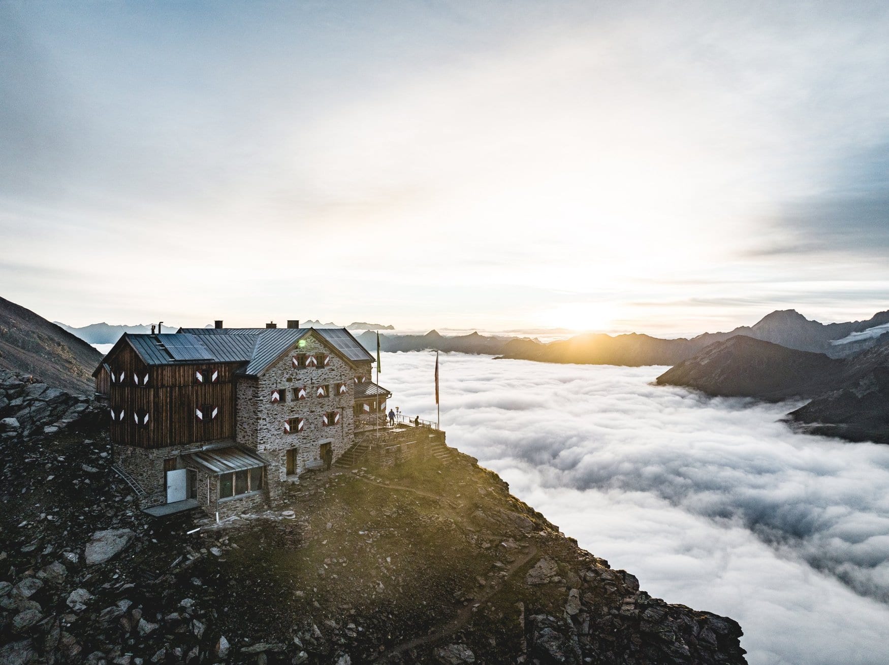 Das Ramolhaus aus Stein und Holz mit rot-weißen Fensterläden steht auf einem Felsvorsprung über einem Nebelmeer, während die Sonne hinter den Bergen aufgeht.