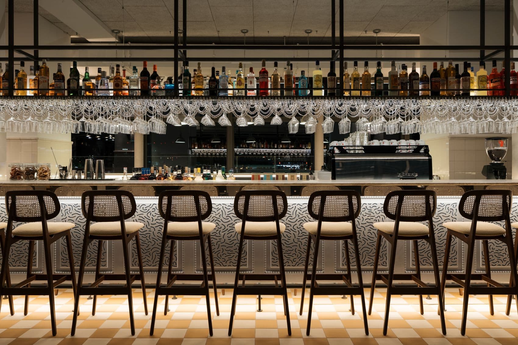 Lilou Bar im Ruby Lilou Marseille mit langer beleuchteter Bartheke, dunklen Holzstühlen, hängenden Weingläsern und Schachbrettboden.
