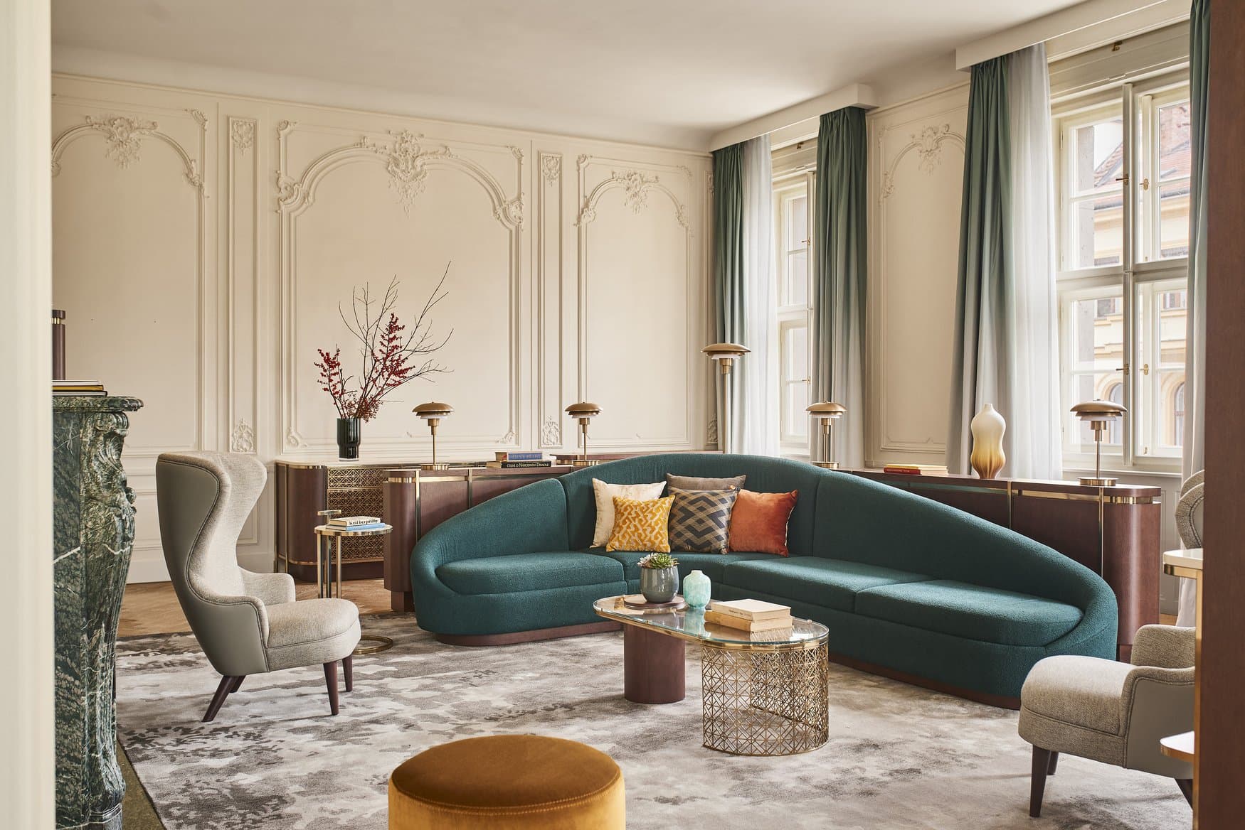 Salon mit Stuckvertäfelung und hohen Fenstern hinter grünen Vorhängen. In der Mitte steht ein geschwungenes türkisfarbenes Sofa mit bunten Kissen, links ein Kamin aus grünem Marmor, davor ein ockerfarbener Hocker.