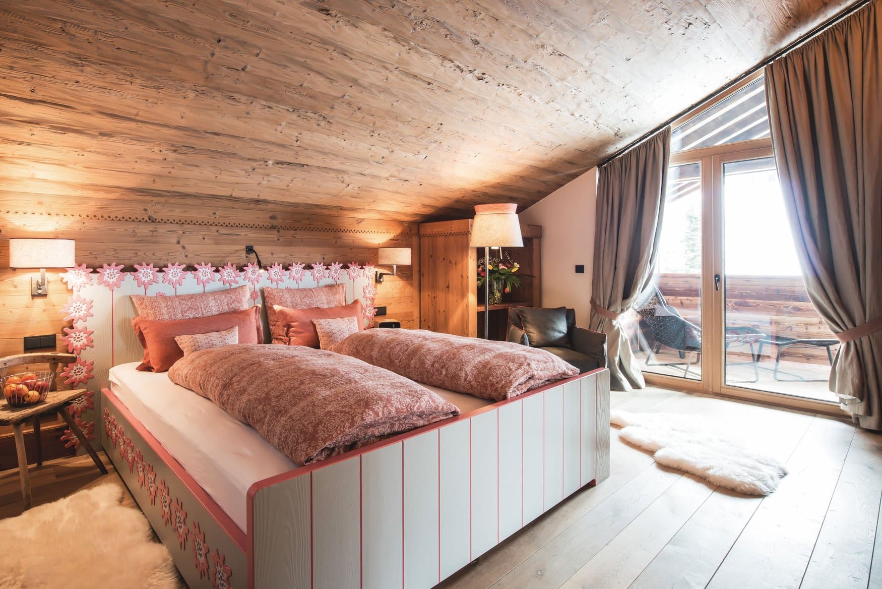 Ein Schlafzimmer unter einer Altholzdecke mit Doppelbett, dessen Kopfteil mit geschnitzten Edelweißblüten besetzt ist. Rechts führt eine Glastür auf den Balkon, auf dem Dielenboden liegen Schaffelle.
