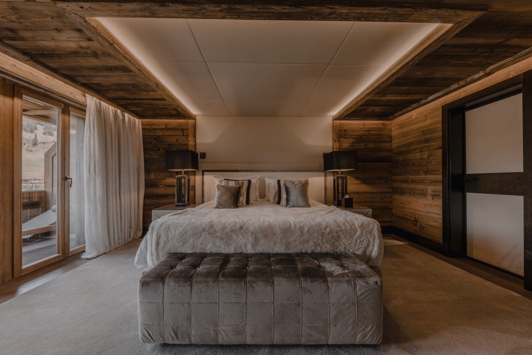 Ein Schlafzimmer mit Altholzverkleidung und abgehängter Lichtdecke. Auf dem Doppelbett liegt ein heller Fellüberwurf, am Fußende steht eine gesteppte Samtbank. Links führt eine Glastür auf einen Balkon mit Liege.